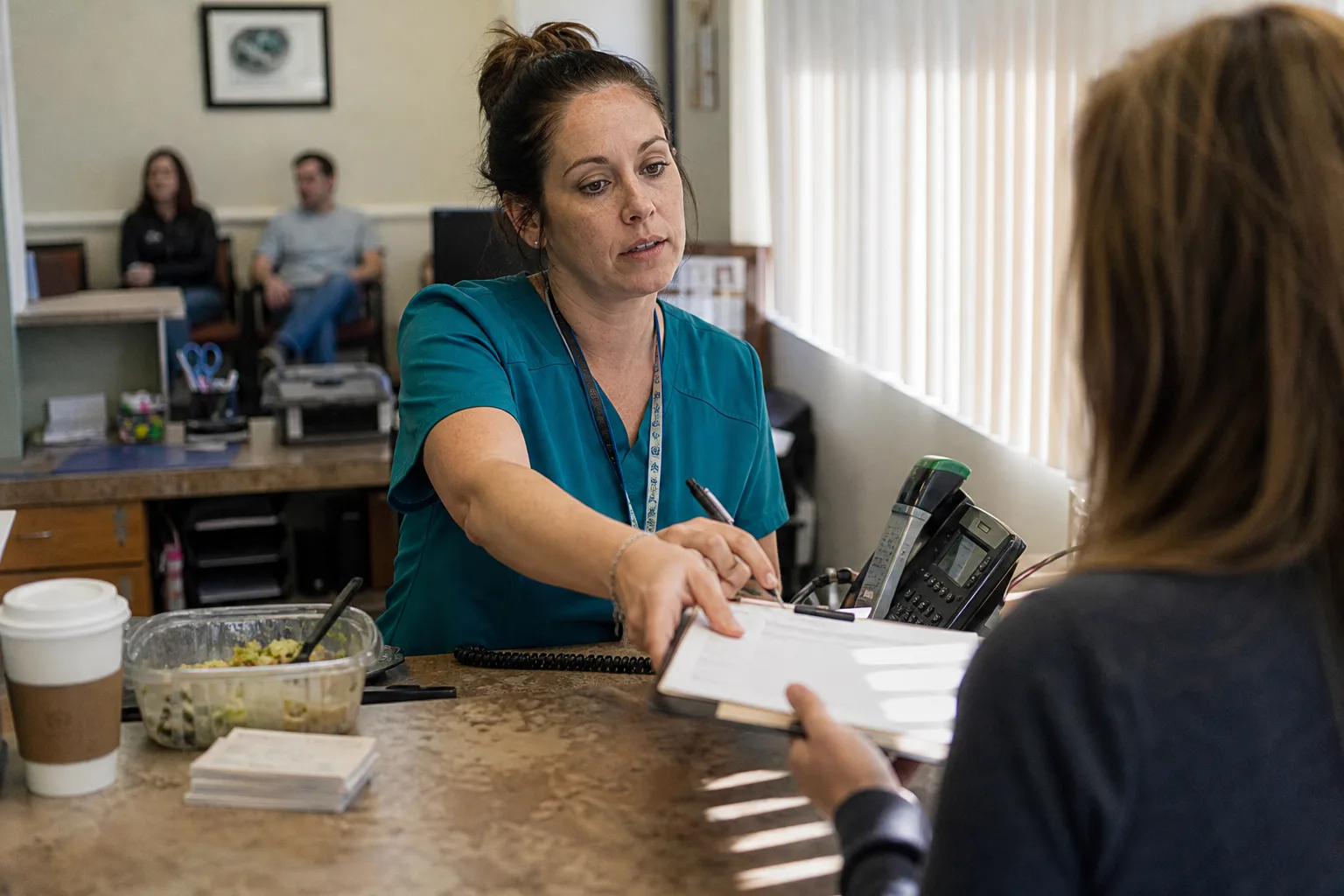 Dental receptionist checking in a patient during lunch rush while a desk phone rings unanswered, the moment missed call recovery matters most