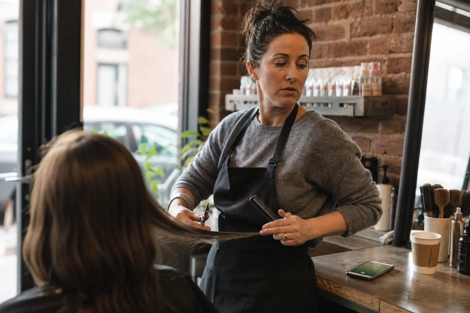 Salon owner in Canada mid-haircut, hands full, glancing at a ringing iPhone she can't answer — the moment an AI phone secretary covers