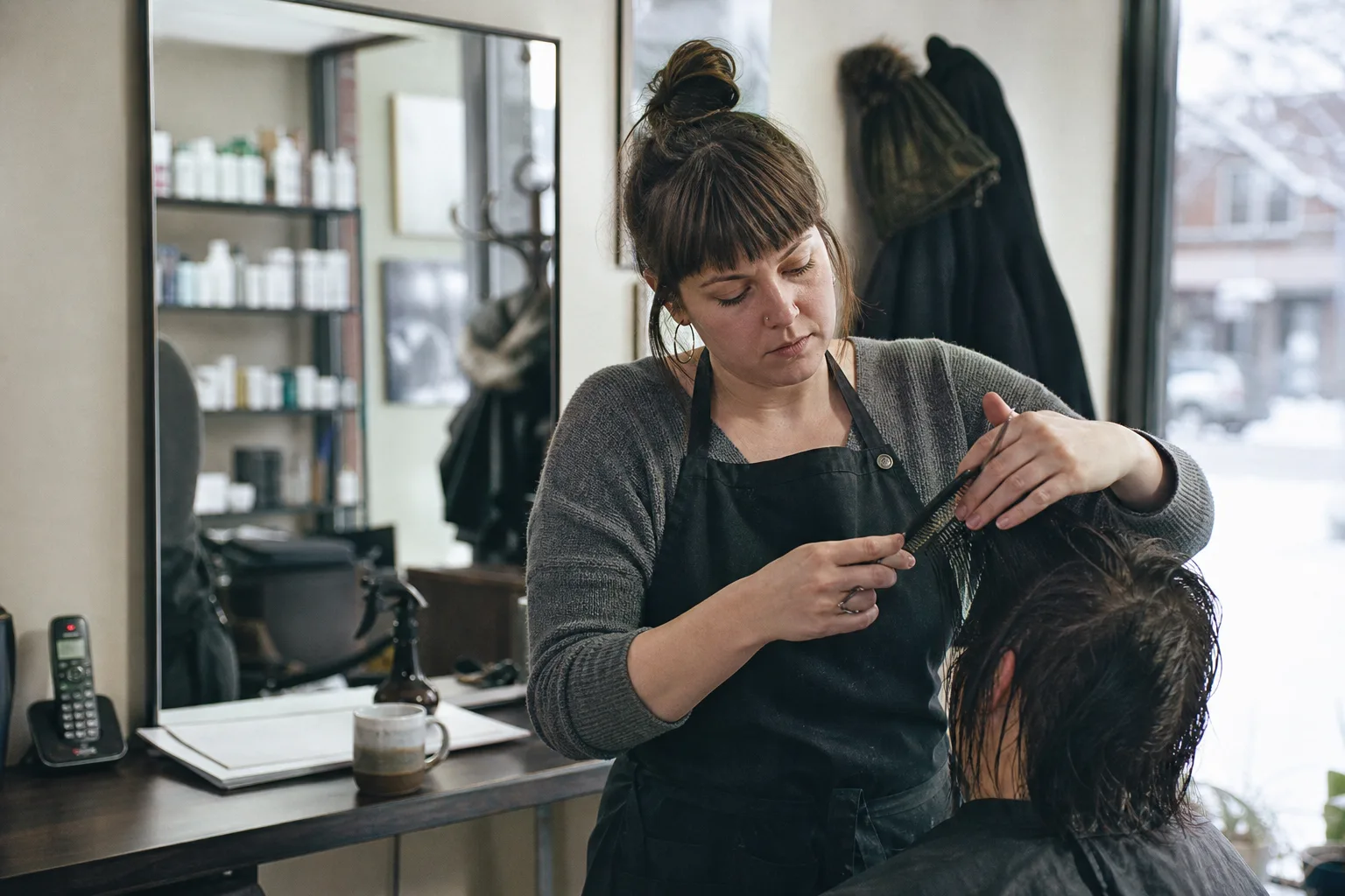 Salon owner cutting a client's hair while the shop phone blinks unanswered on the counter, the routine call an ai receptionist would take