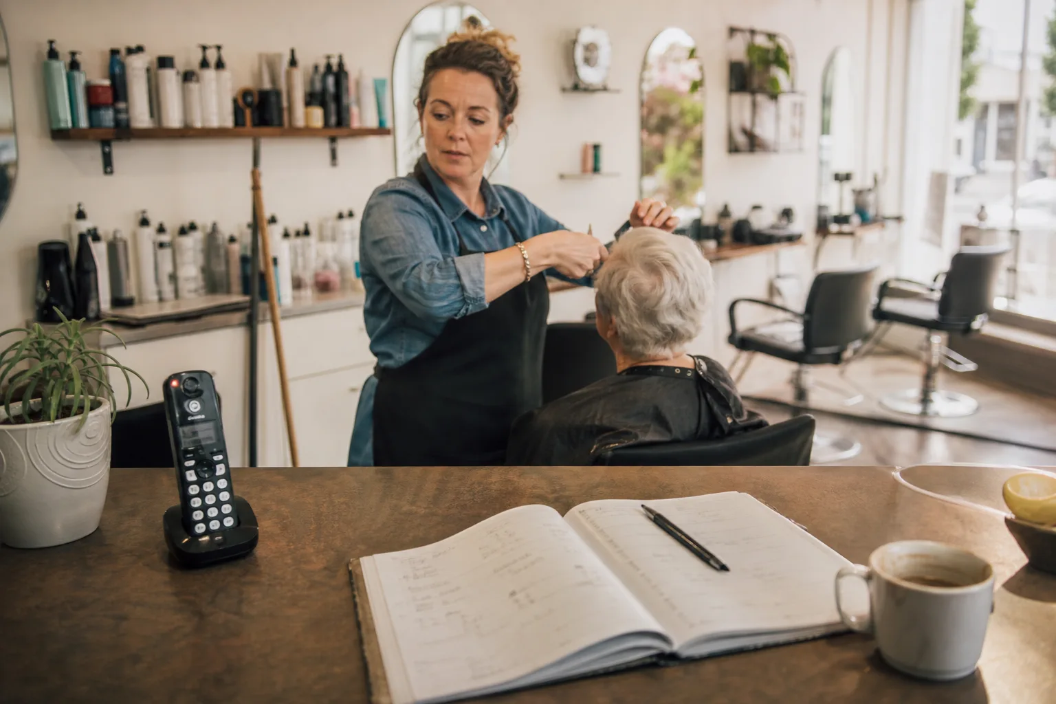 Salon owner in Christchurch cutting a client's hair while the front desk phone rings unanswered beside an open appointment diary